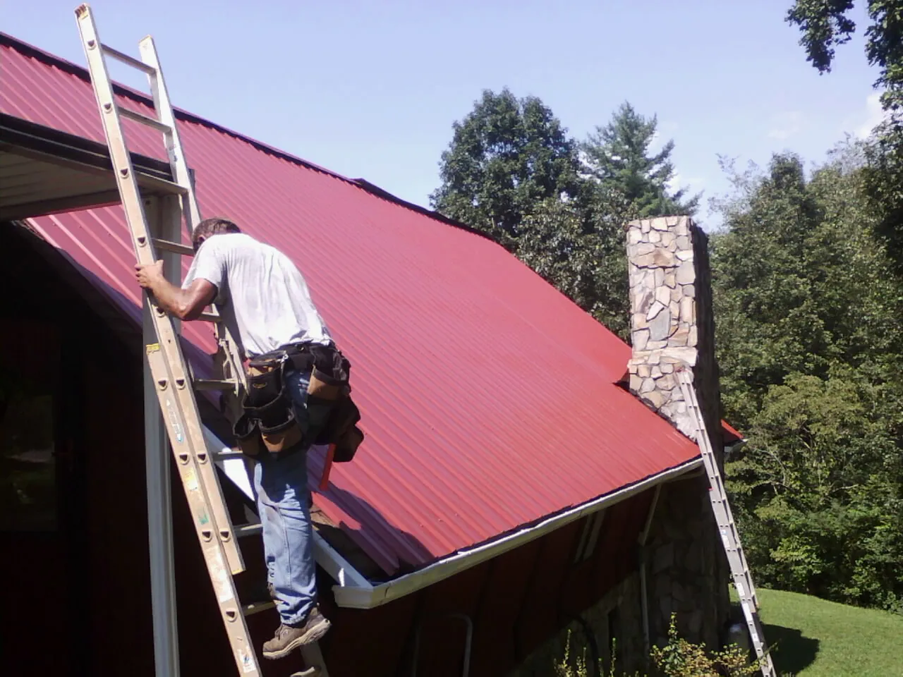 Roofer on a ladder beside a red metal roof and stone chimney — Soffit & Fascia Repair by Roofing Brothers in Marshallville